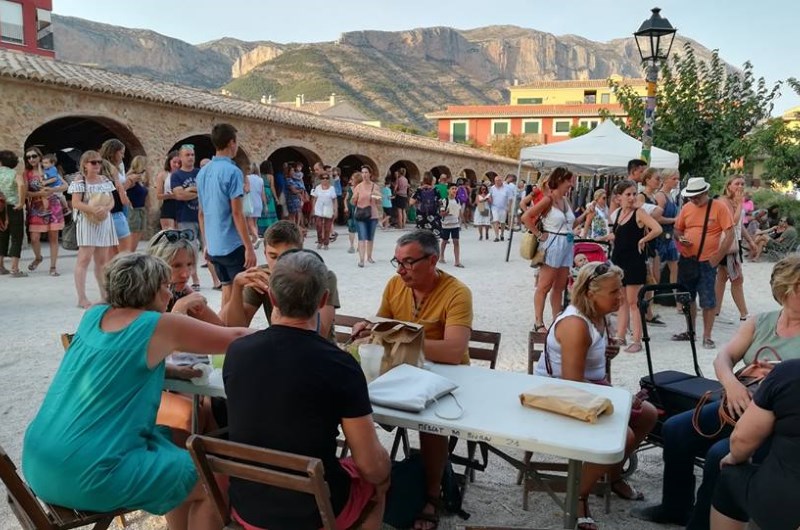 Foto en la que se ve a gente en un mesa disfrutando del mercado con el Riurau de fondo.